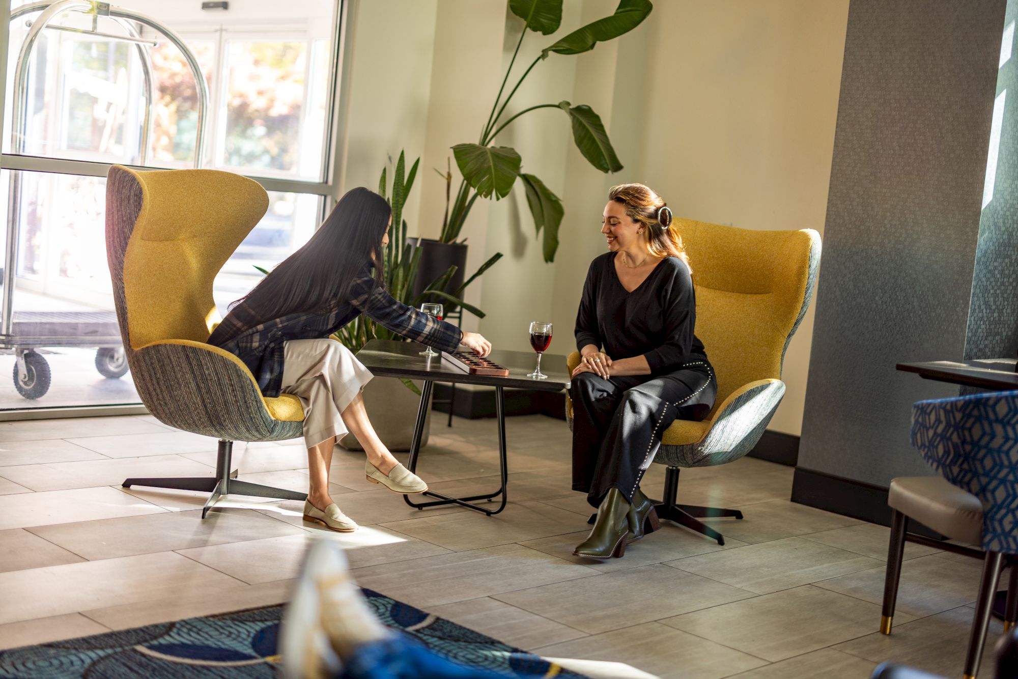 Two people relax in a stylish lounge, one in a yellow chair with legs up, the other seated calmly with drinks on a small table, nearby plants.