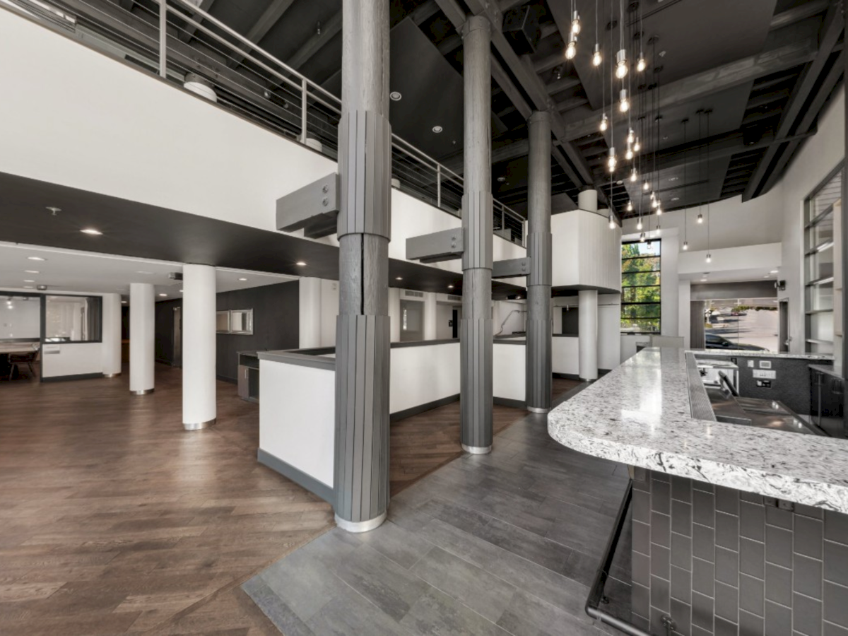 Modern office kitchen/lounge with a granite island, dark cabinets, exposed beams, tall columns, pendant lights, and wood floors.