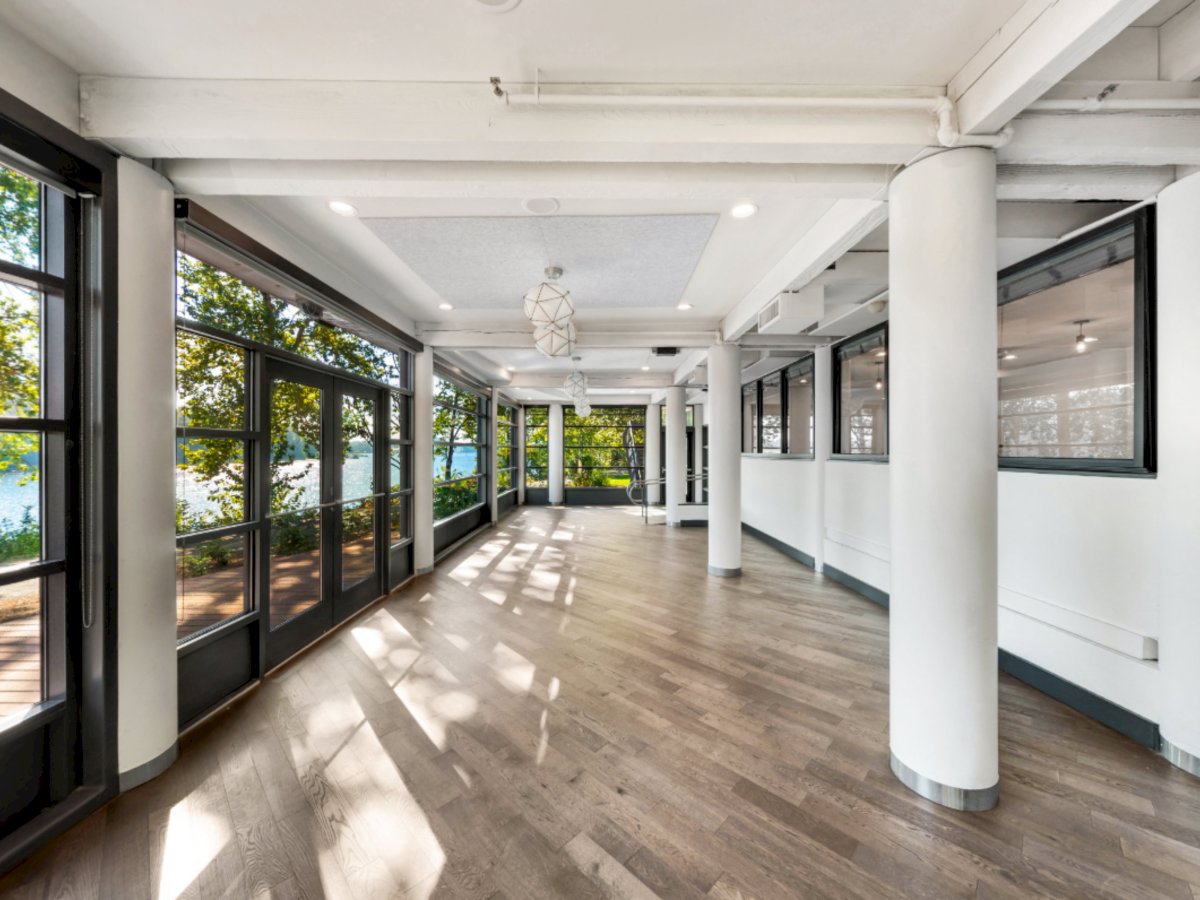 A bright modern hallway with large floor-to-ceiling windows, white columns, and wooden flooring stretching toward a seating area, bathed in sunlight.