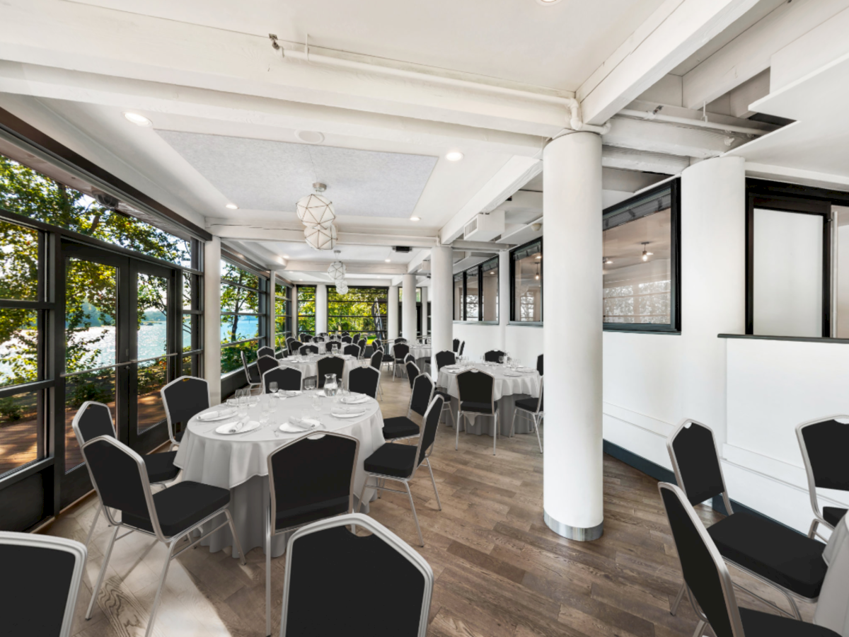 A modern lakeside banquet hall with round tables, black chairs, white decor, and large windows letting in daylight.
