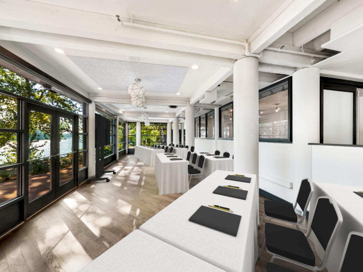 A bright meeting room with long tables, black chairs, pendant lights, and large windows beside greenery and a lake.