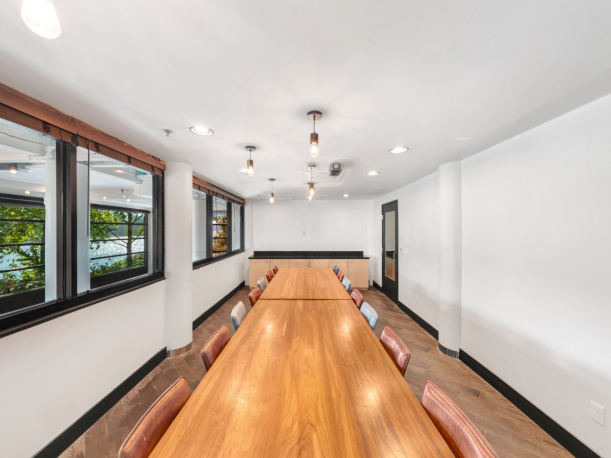 Bright meeting room with a long wooden table, leather chairs, pendant lights, and big windows with trees outside.
