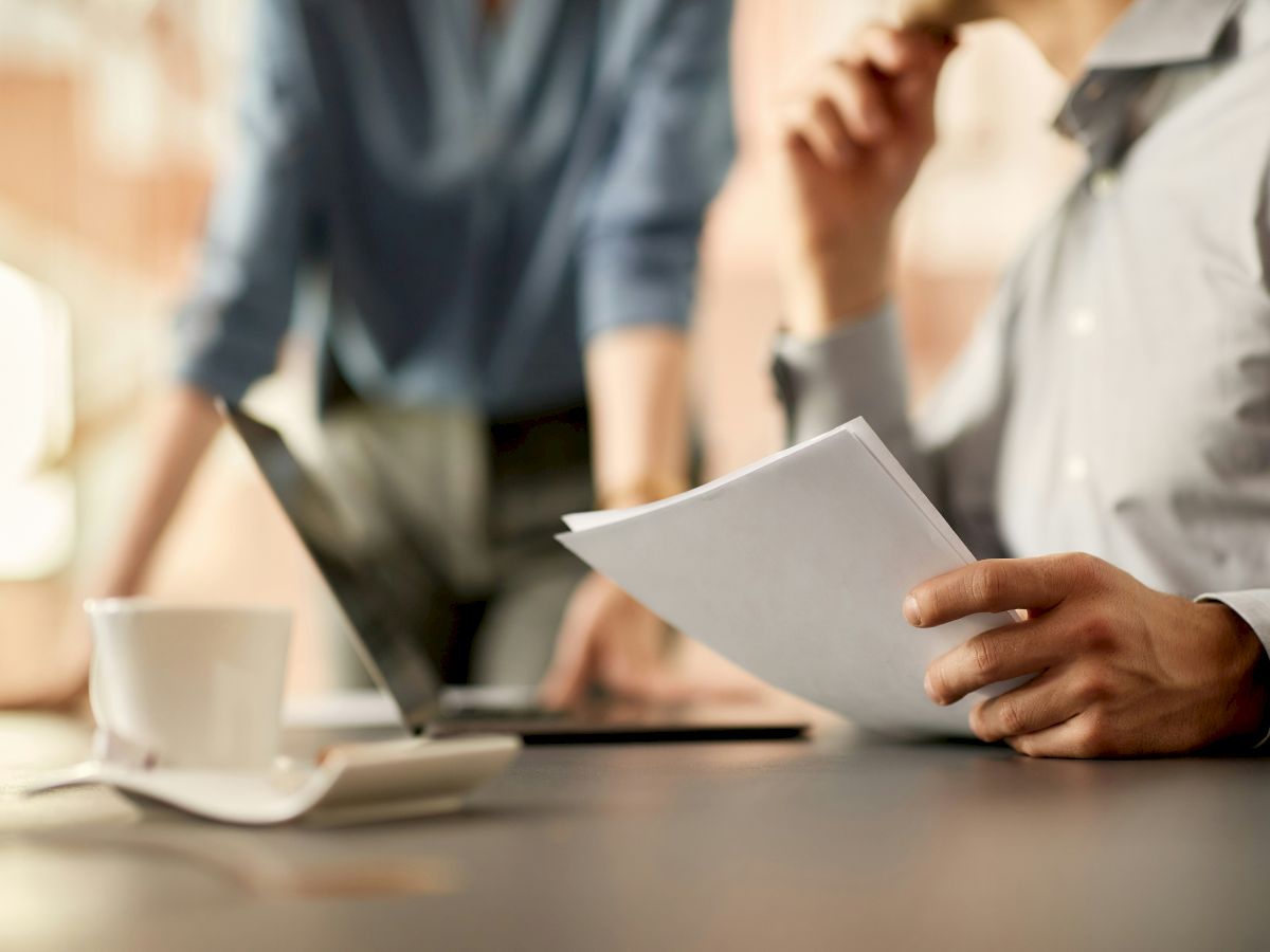 Two people are discussing in an office setting. One holds papers, the other leans over a laptop. A coffee cup is on the table.