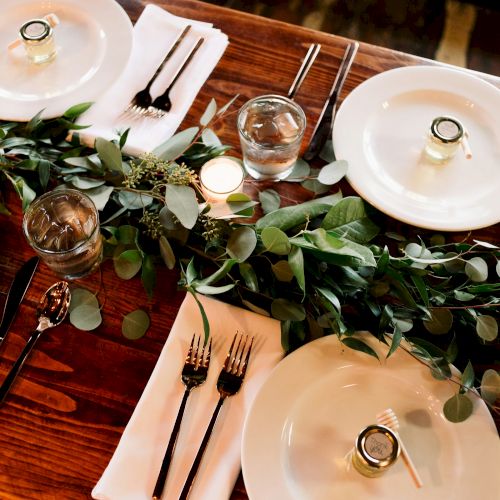 A wooden table is elegantly set with white plates, cutlery, glasses, candles, and a green foliage centerpiece, creating a cozy dining atmosphere.