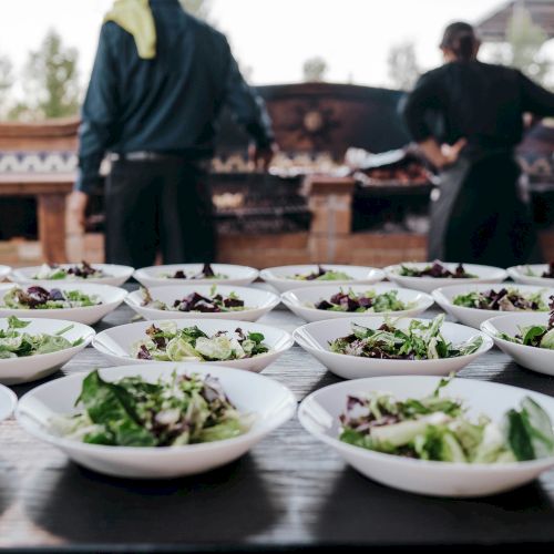 A table with bowls of salad is prepared, while chefs work in the background of an outdoor setting.