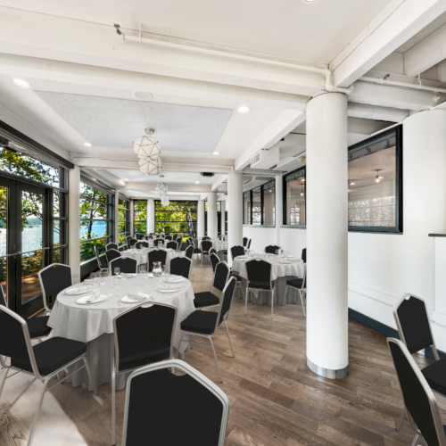 A bright event room with round tables set for a banquet, black chairs, large windows, and views of trees and a lake.