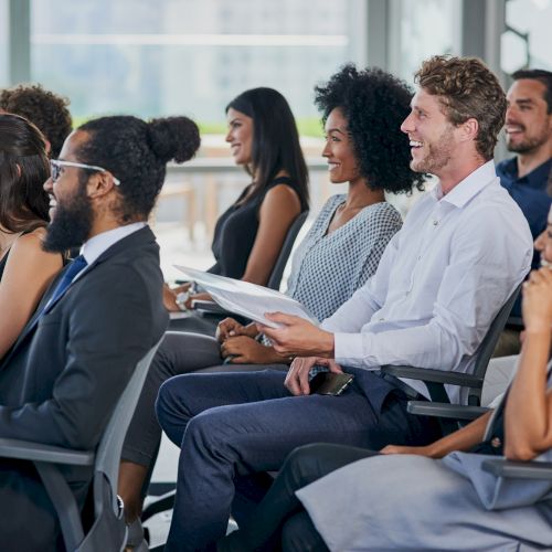 A diverse group of people seated in chairs, smiling and engaging in what appears to be a meeting or lecture in a bright room.