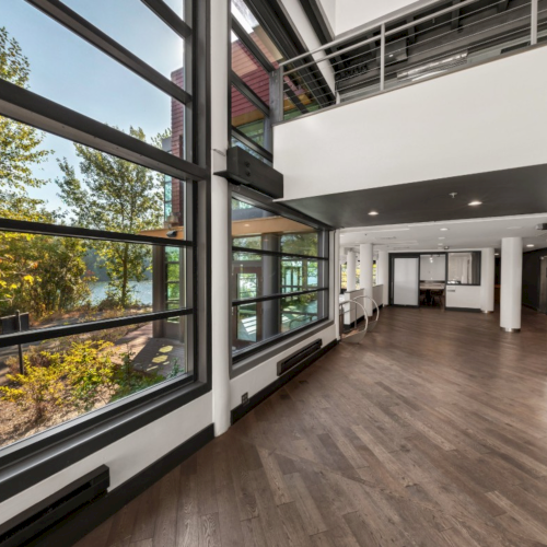 Bright modern indoor corridor with large windows and wooden floors, overlooking trees and a driveway.