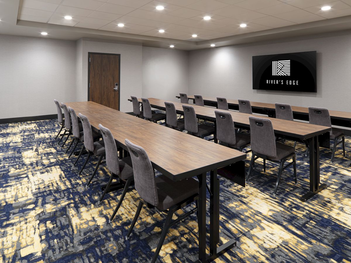 A modern conference room with long wooden tables, gray chairs, carpeted floor, a wall-mounted screen at the front, and bright ceiling lights.
