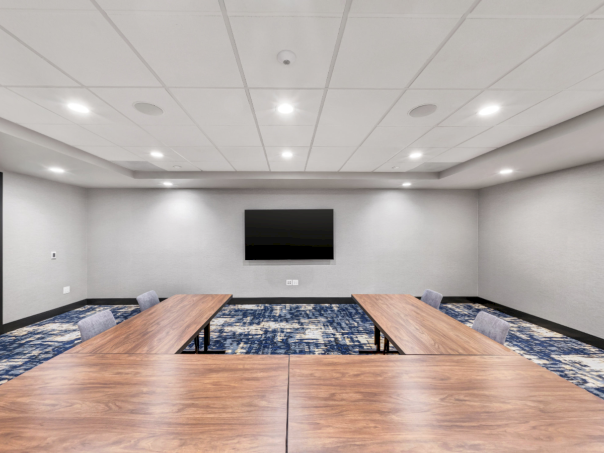 A modern conference room with two wooden tables, blue patterned carpet, ceiling lights, and a wall-mounted TV.
