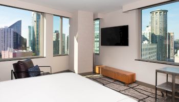 The image shows a modern hotel room with large windows, city views, a bed, TV, armchair, and minimalist decor.