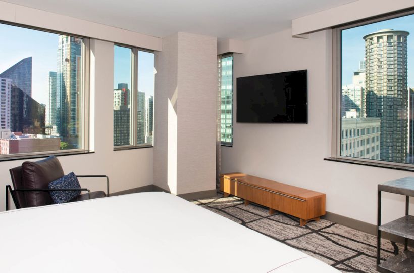The image shows a modern hotel room with large windows, city views, a bed, TV, armchair, and minimalist decor.