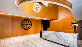 A modern reception area with wooden panels, a large clock, a marble desk, and decorative lighting and flowers.