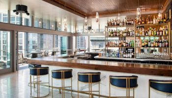 The image shows a modern bar with a wooden ceiling, high chairs, and a well-stocked selection of spirits, overlooking city skyscrapers.