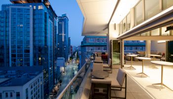 A modern rooftop terrace overlooking a cityscape at dusk, with seating areas and adjacent glass-fronted buildings illuminated by city lights.