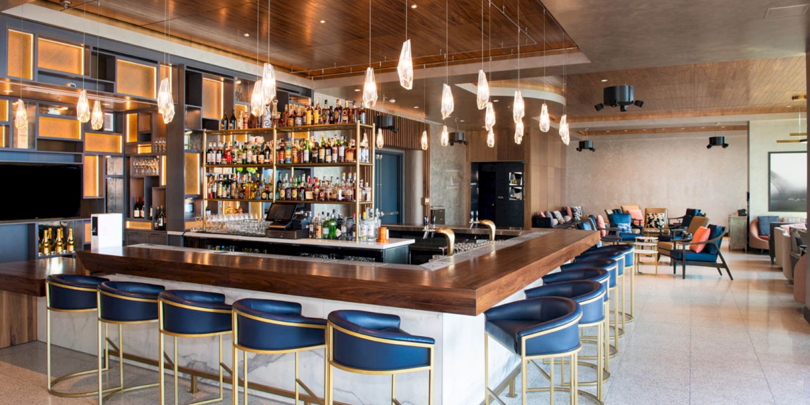 The image shows a modern bar with blue stools, hanging lights, and a well-stocked counter, in a spacious interior setting.
