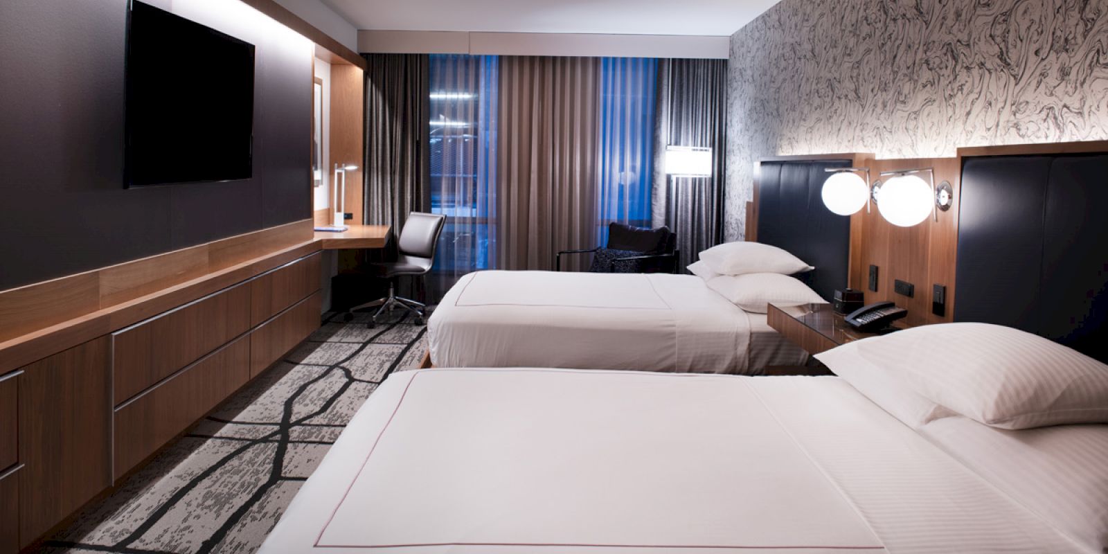 A hotel room with two beds, a desk and chair, a wall-mounted TV, and a window with curtains, lit by modern lamps on the wall.