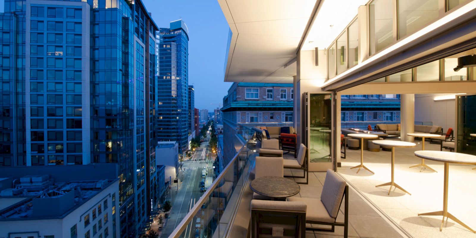 A modern rooftop terrace overlooking a cityscape at dusk, with seating areas and tall buildings lit up in the background.