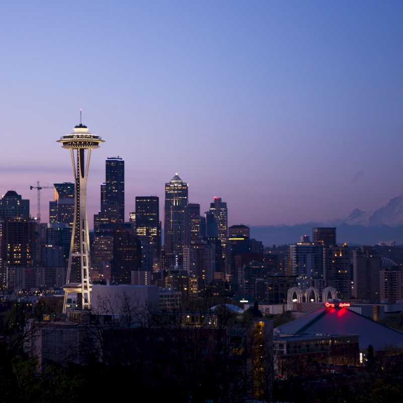 Seattle skyline featuring the Space Needle at dusk, with a view of Mount Rainier in the background under a purple sky.