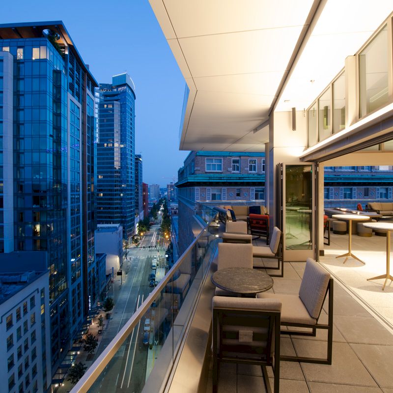 The image shows a modern outdoor terrace with seating, overlooking a city street and tall buildings during the evening.