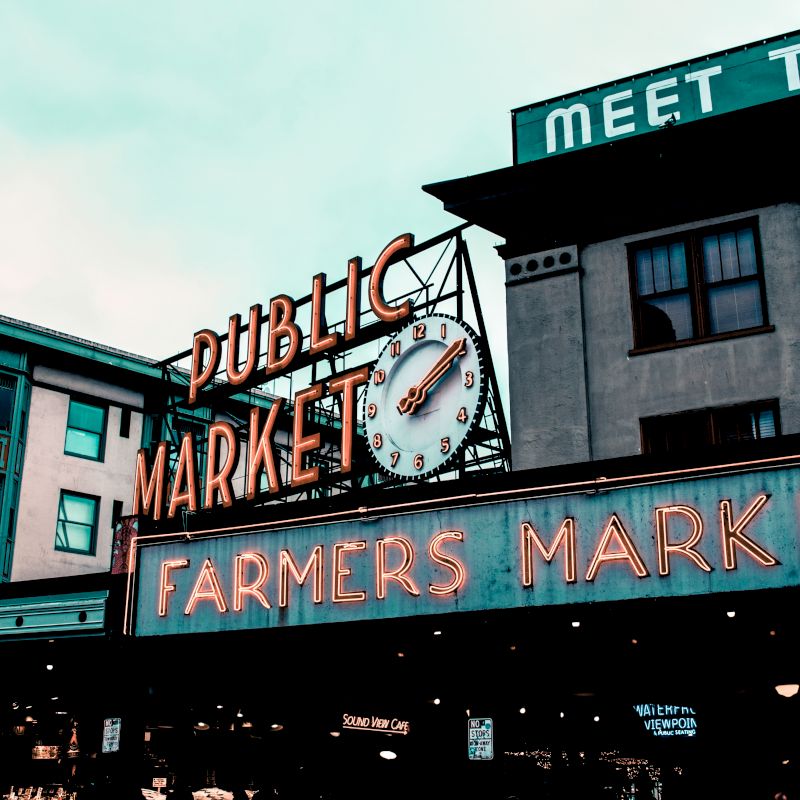 The image shows a neon sign for a public farmers market, with a clock above, set against a backdrop of urban buildings.