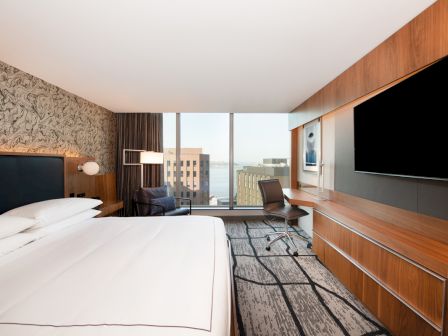 A modern hotel room with a large bed, flat-screen TV, desk area, and city view through a large window, creating a cozy atmosphere.