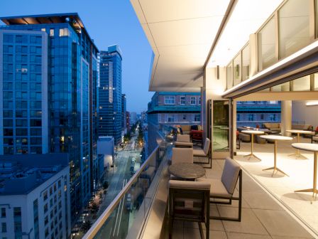 A modern rooftop terrace with seating areas overlooks a cityscape of tall buildings and a street below, with lights illuminating the evening scene.