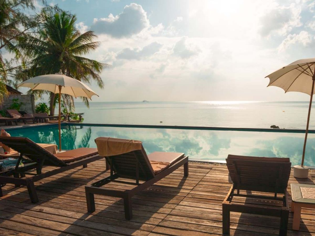 A serene beachside view with lounge chairs, umbrellas, palm trees, and a calm sea under a partly cloudy sky.