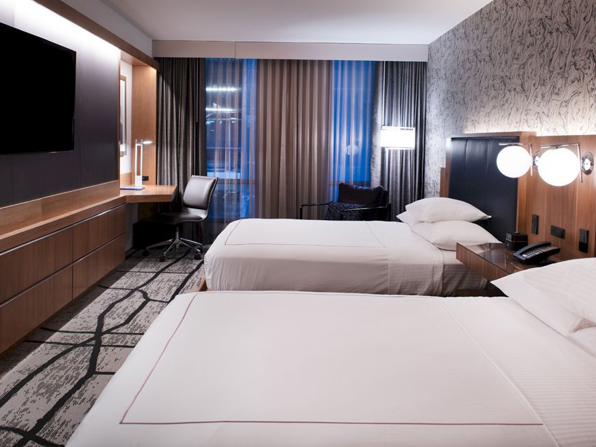 The image shows a modern hotel room with two beds, a wall-mounted TV, a desk, lamps, and a window with curtains, all in a stylish decor.