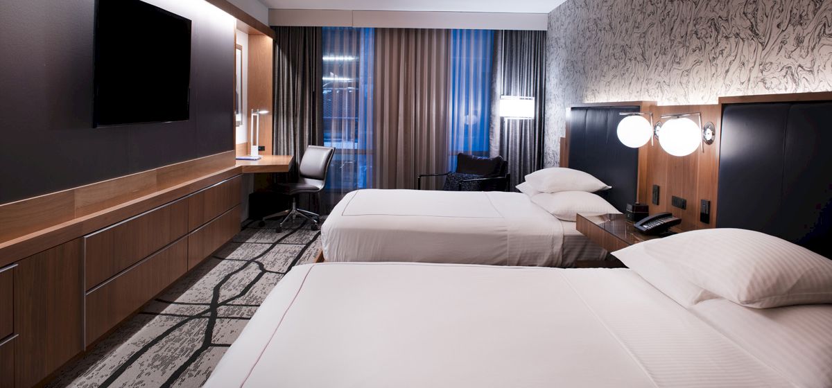 The image shows a modern hotel room with two beds, a wall-mounted TV, a desk, lamps, and a window with curtains, all in a stylish decor.
