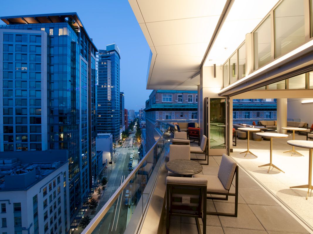 A modern cityscape at dusk with illuminated buildings and a contemporary outdoor patio setting above, featuring seating and tables.