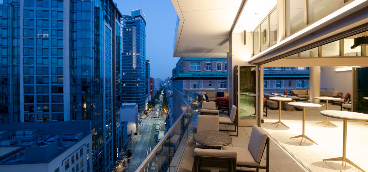 A modern cityscape at dusk with illuminated buildings and a contemporary outdoor patio setting above, featuring seating and tables.