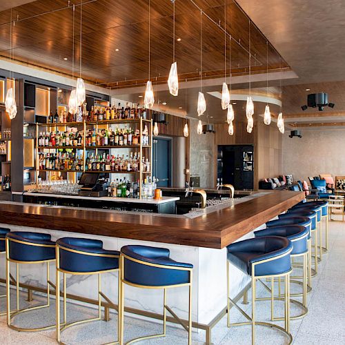 The image shows a modern bar with a central counter, blue stools, and pendant lights, creating a stylish and inviting atmosphere.
