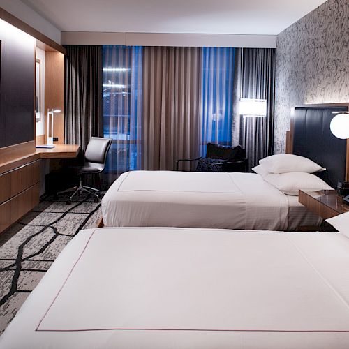 A hotel room with two beds, a TV, a desk, and large window with curtains. The room has modern decor and ambient lighting.