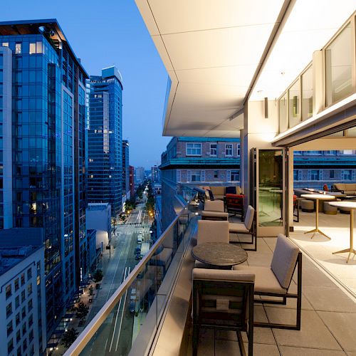 A modern urban rooftop terrace is overlooking a city with tall buildings at dusk, featuring outdoor seating and glass walls.