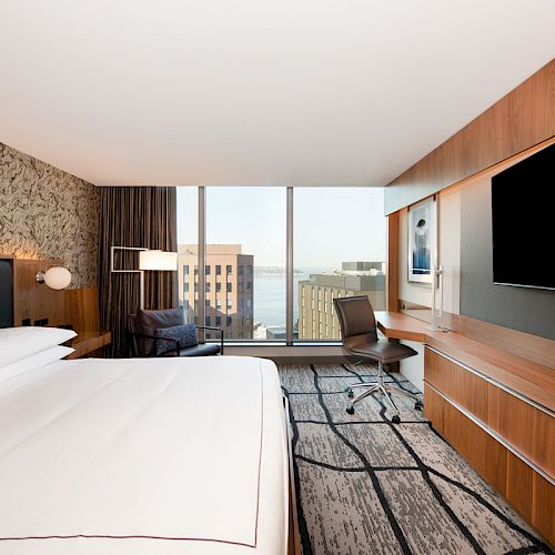 A modern hotel room with a large bed, flat-screen TV, desk, chair, and a window offering a view of buildings outside.