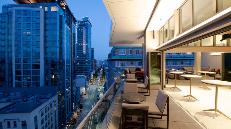 A modern rooftop terrace at dusk with seating areas, overlooking a cityscape of tall buildings and illuminated streets.