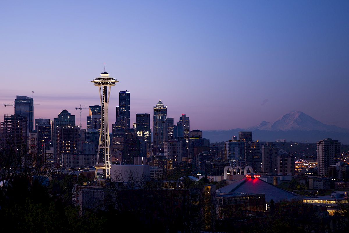 The image shows Seattle's skyline at dusk, featuring the Space Needle and skyscrapers, with a mountain in the background under a twilight sky.