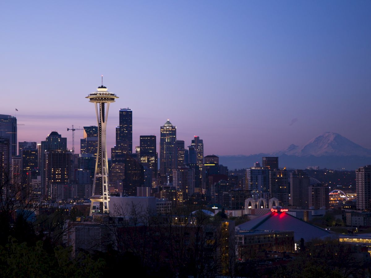 The image shows Seattle's skyline at dusk, featuring the Space Needle and skyscrapers, with a mountain in the background under a twilight sky.