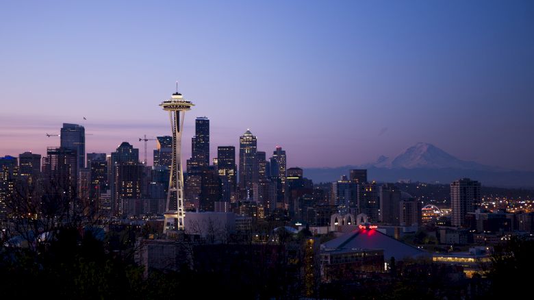 The image shows Seattle's skyline at dusk, featuring the Space Needle and skyscrapers, with a mountain in the background under a twilight sky.