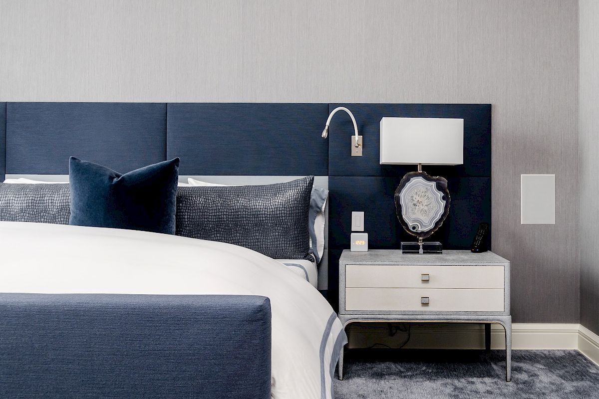 A modern bedroom with a blue headboard, white nightstand, lamp, and a decorative pillow on the bed.