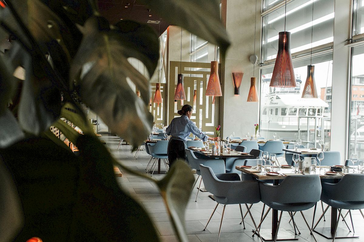 A person is arranging tables in a modern, elegant restaurant with large windows, contemporary lighting, and stylish furniture.