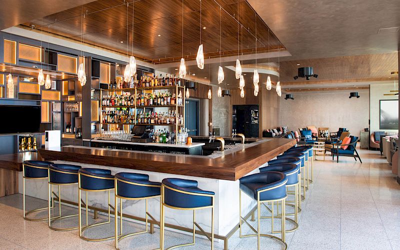 The image shows a modern bar with blue stools, a wooden countertop, pendant lights, and shelves stocked with bottles.