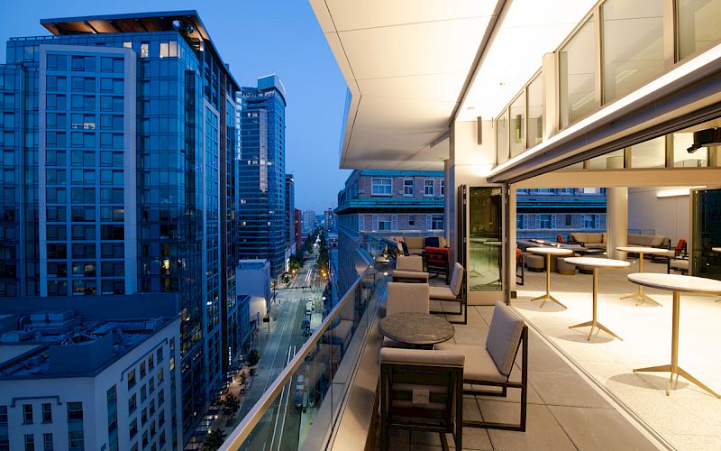 A modern rooftop terrace at dusk with seating, overlooking a cityscape of tall buildings and a lit street.