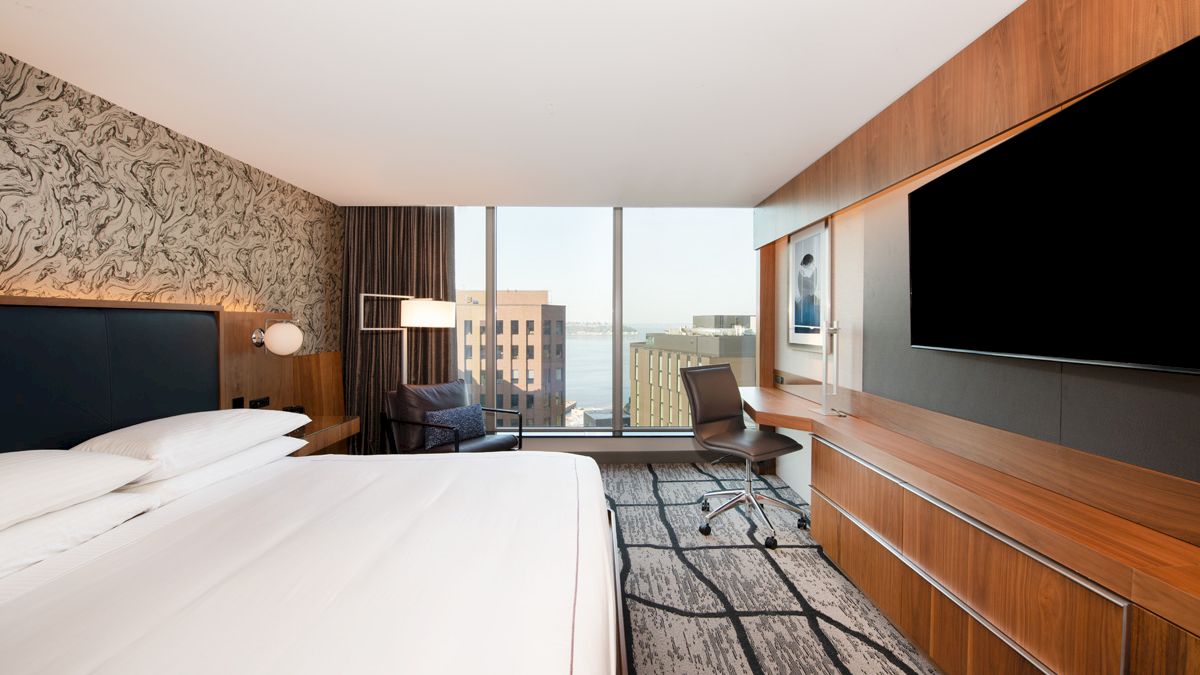 A modern hotel room features a large bed, TV, desk with chair, and floor-to-ceiling window offering a city view.