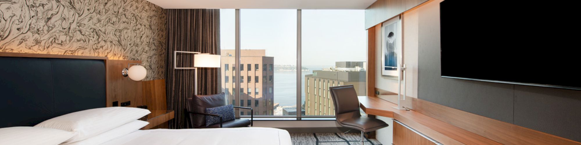 A modern hotel room features a large bed, TV, desk with chair, and floor-to-ceiling window offering a city view.